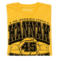 a folded gold shirt with black print that says, "The Incredible Stuelk. Hannah Stuelke. Forward. Cedar Rapids" over an illustration of a giant basketball and a basketball court.