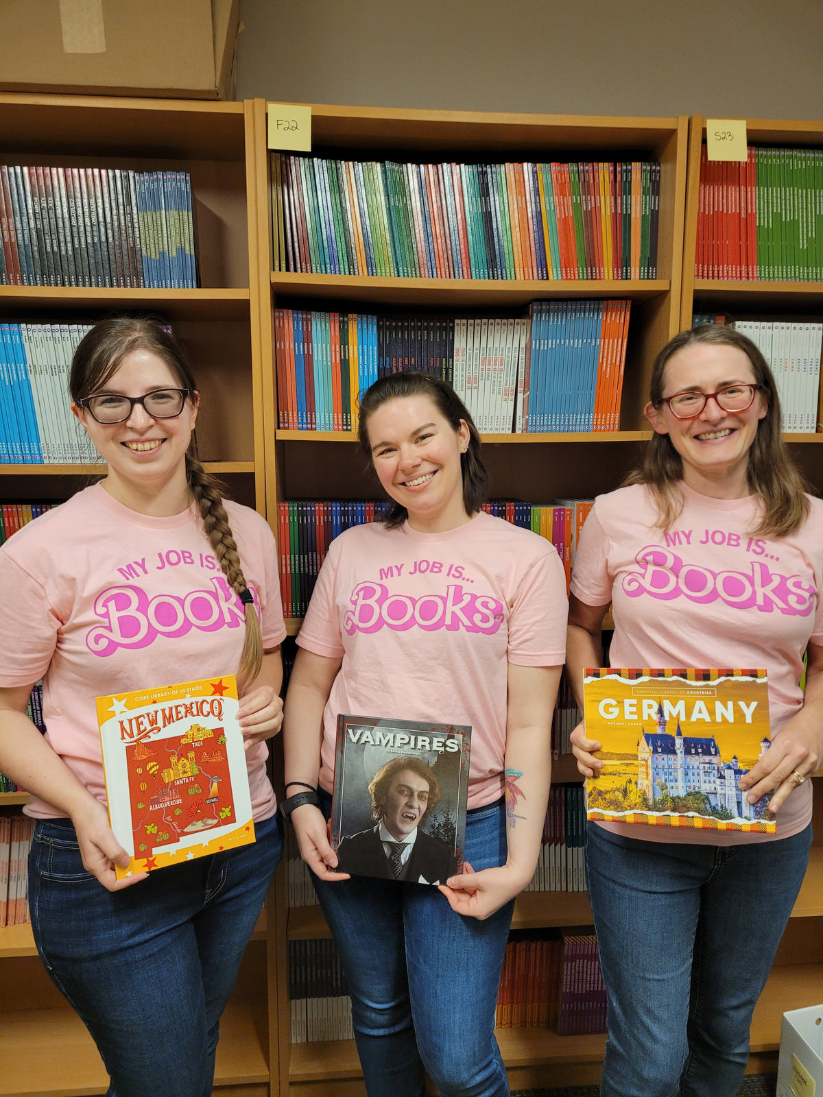 Three women wearing union made My Job Is... Books t-shirts stand in front of a bookshelf, each holding a different book about New Mexico, vampires, and Germany.