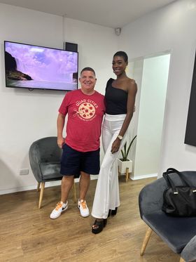 A man in a red RAYGUN Quad Cities t-shirt and shorts stands next to a tall woman wearing a black one-shoulder top and white high-waisted pants.