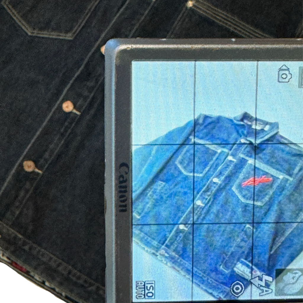 A digital camera screen displaying a photo of a blue vintage Kanijeans jacket with a red logo on the chest, positioned over a dark wash denim jacket.