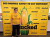 A woman wearing a Heather Mellow Yellow t-shirt that says Sweet like corn stands in front of a yellow promotional backdrop for the musical Shucked at the Des Moines Civic Center.