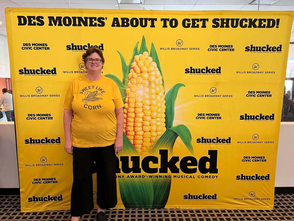 A woman wearing a Heather Mellow Yellow t-shirt that says Sweet like corn stands in front of a yellow promotional backdrop for the musical Shucked at the Des Moines Civic Center.