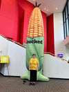 A woman wearing a Heather Mellow Yellow t-shirt stands in front of a giant inflatable ear of corn advertising the musical Shucked in a building lobby.