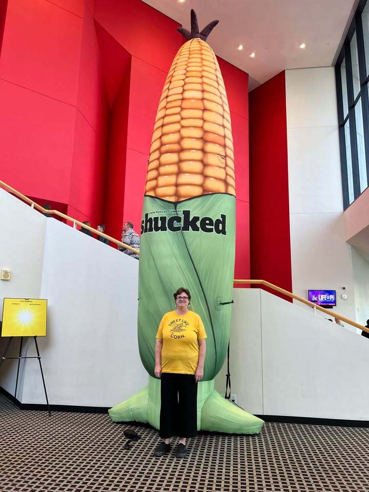 A woman wearing a Heather Mellow Yellow t-shirt stands in front of a giant inflatable ear of corn advertising the musical Shucked in a building lobby.
