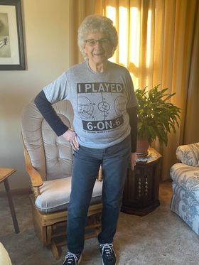 An older woman with short gray hair wears a heather grey RAYGUN T-shirt that reads I Played 6-on-6 over a black long-sleeved shirt, standing in a room with a wooden rocking chair.