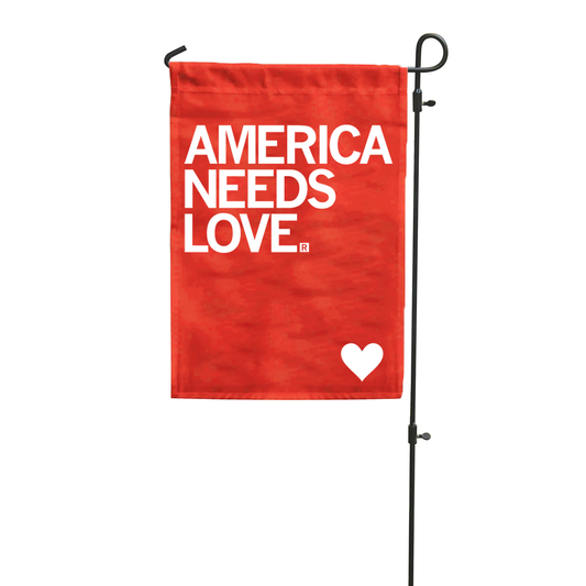 A red polyester garden flag on a black metal stand displays the white text AMERICA NEEDS LOVE with a small white heart at the bottom.