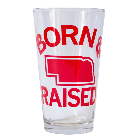 Clear dishwasher-safe pint glass printed with the words BORN & RAISED in red, featuring a red silhouette of the state of Nebraska between the lines of text.