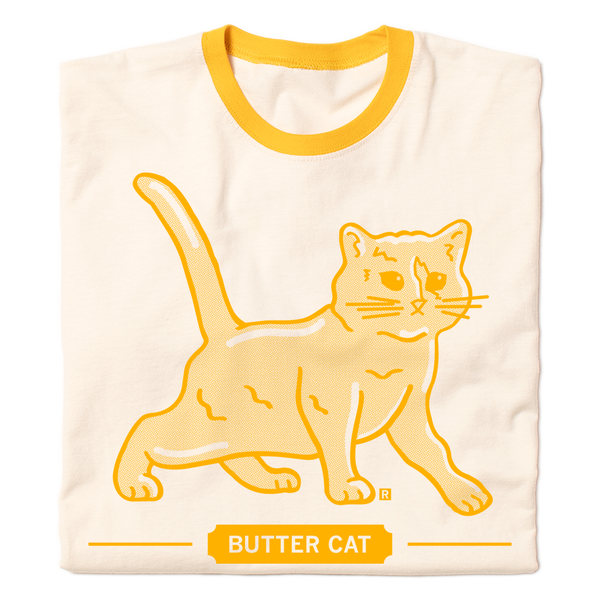 A folded vintage white ringer shirt with yellow sleeve and collar accents. Gold ink printed with a gradient depicts a cat made of butter, spoofing the butter cow. Text below says 