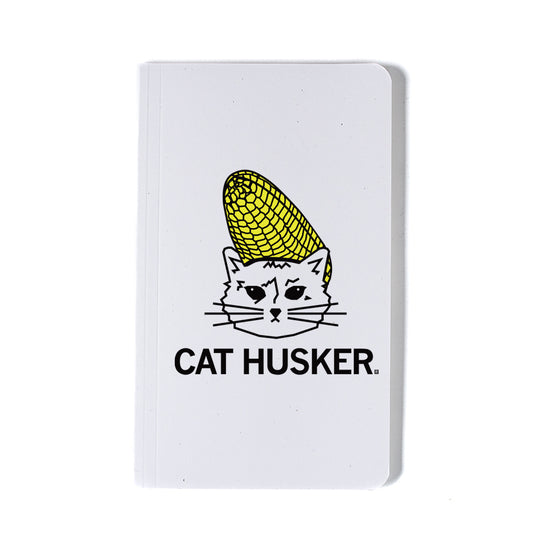 White notebook featuring a screen print of a cat wearing a corn cob as a hat, with the text CAT HUSKER printed below.