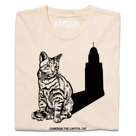 Cream-colored Cameron the Capitol Cat t-shirt featuring a line drawing of a cat sitting in front of a silhouette of the Iowa State Capitol building, with the text CAMERON THE CAPITOL CAT printed below.