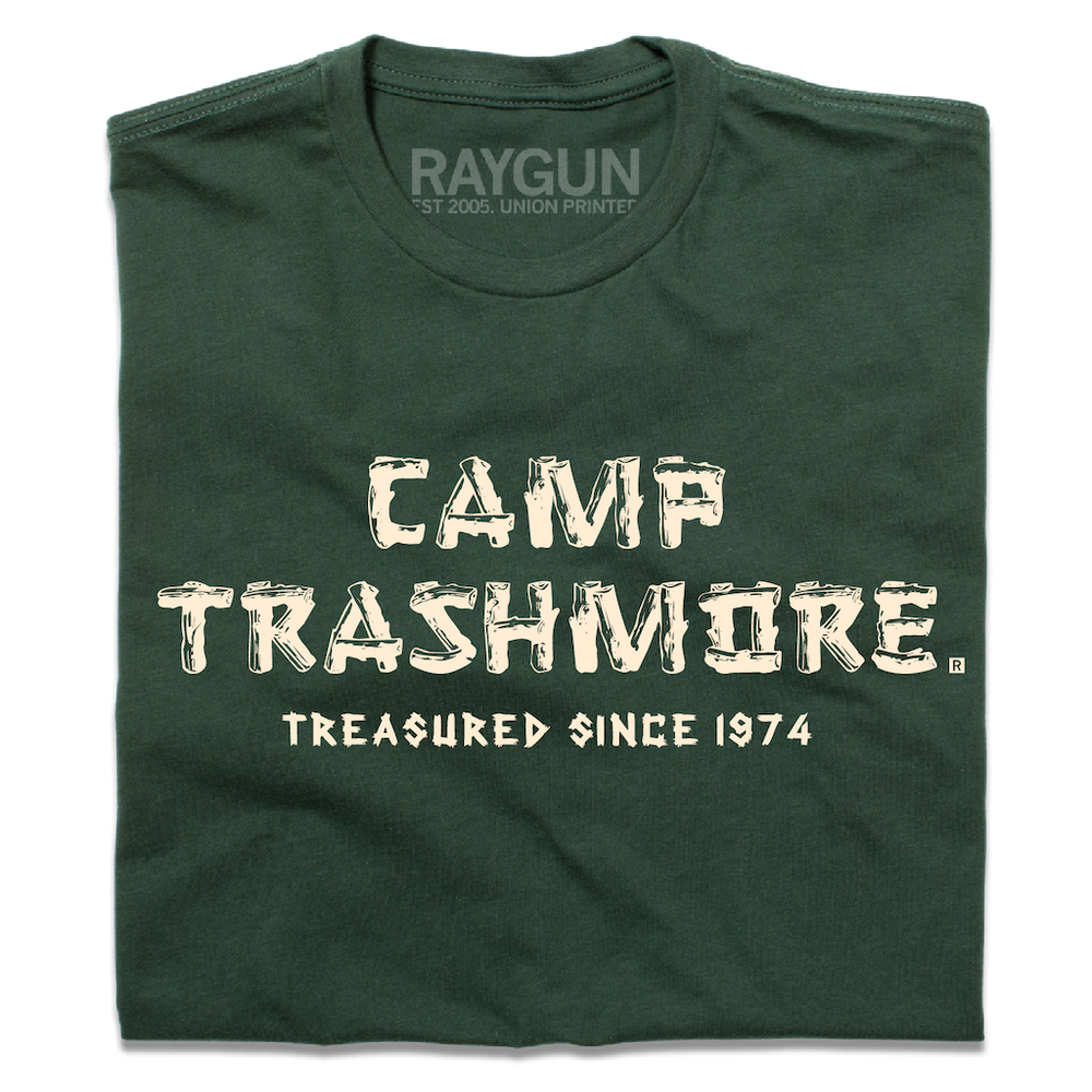 A folded forest green t-shirt with off-white ink that says, "Camp Trashmore. Treasured since 1974." printed in a font reminiscent of old summer camps.