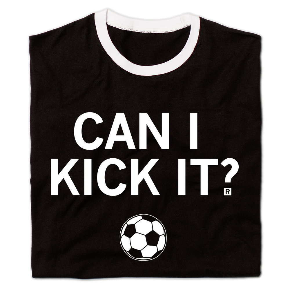 A folded black ringer shirt with a white neckline and sleeves. Centered white ink text reads can I kick it?" above an illustration of a soccer ball.