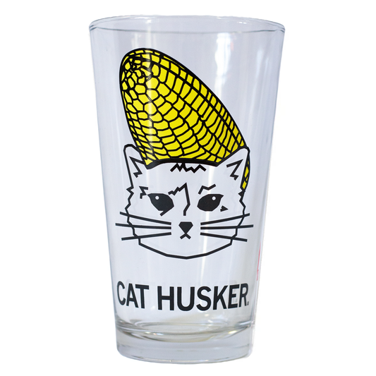 A clear pint glass featuring a line drawing of a cat wearing a corn cob like a hat, with the text CAT HUSKER printed below in soy-based ink.