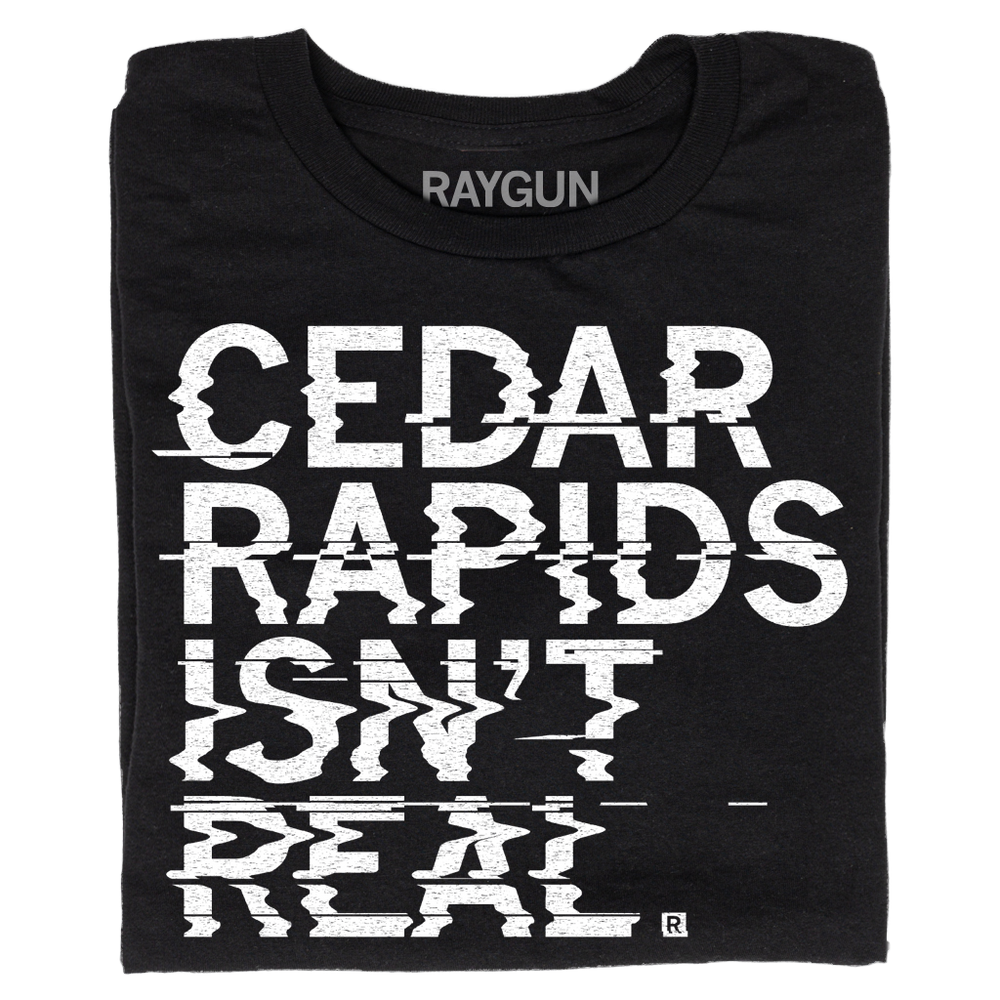 A folded black t-shirt with left-aligned white ink text printed in a distorted style that reads "Cedar Rapids Isn't Real"