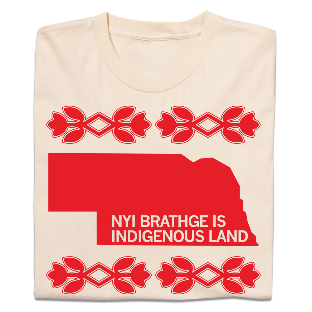 A folded cream t-shirt with bright red ink print of Nebraska with cream text over the bottom of the state that says, "NYI BRATHGE IS INDIGENOUS LAND". 