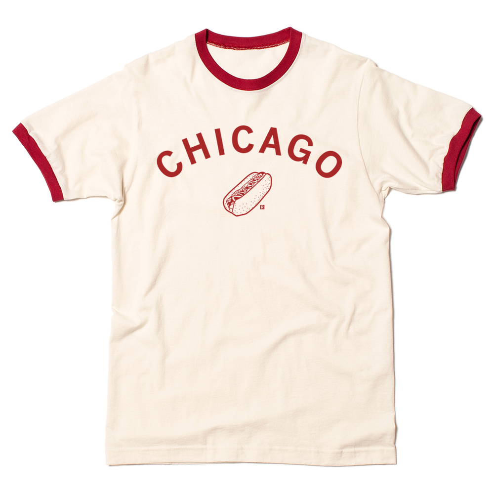 A folded ivory and red ringer shirt with  curved red ink text that says, "Chicago" over an illustration of a Chicago style hot dog.