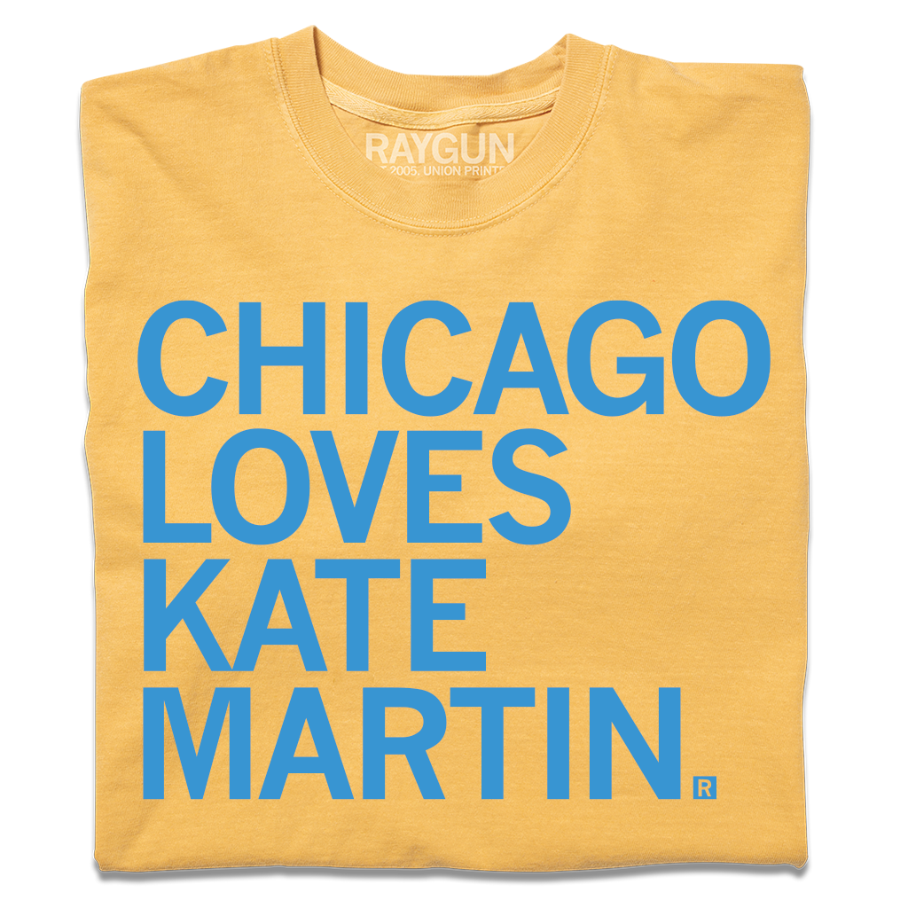 A folded heavyweight mustard t-shirt with left-aligned bright blue text that reads "CHICAGO LOVES KATE MARTIN."
