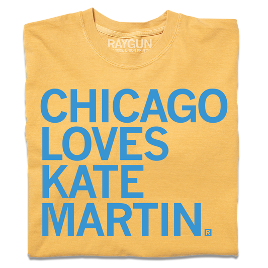 A folded heavyweight mustard t-shirt with left-aligned bright blue text that reads "CHICAGO LOVES KATE MARTIN."
