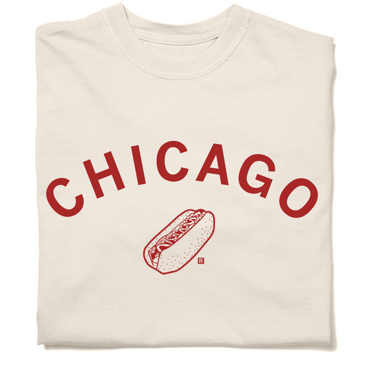 A folded off white t-shirt with curved red ink text that says, "Chicago" over an illustration of a Chicago style hot dog.