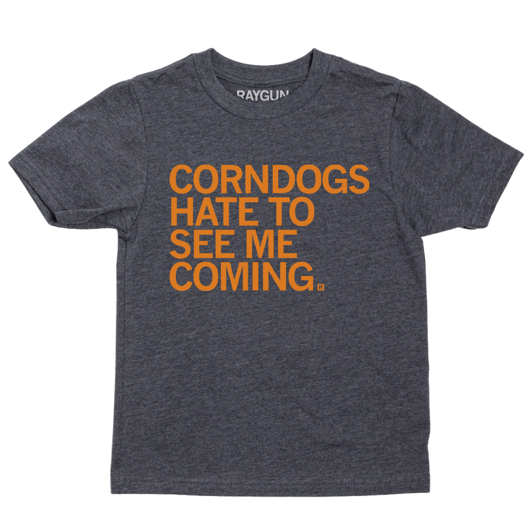 A youth size heather black t-shirt with orange print text that says, "CORNDOGS HATE TO SEE ME COMING"