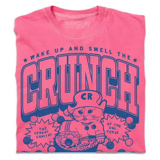 Pink heavyweight cotton t-shirt with a blue graphic of a cat wearing a cereal bowl hat above the text WAKE UP AND SMELL THE CRUNCH, THE CEREAL CAPITAL, and OF THE UNI-VERSE.