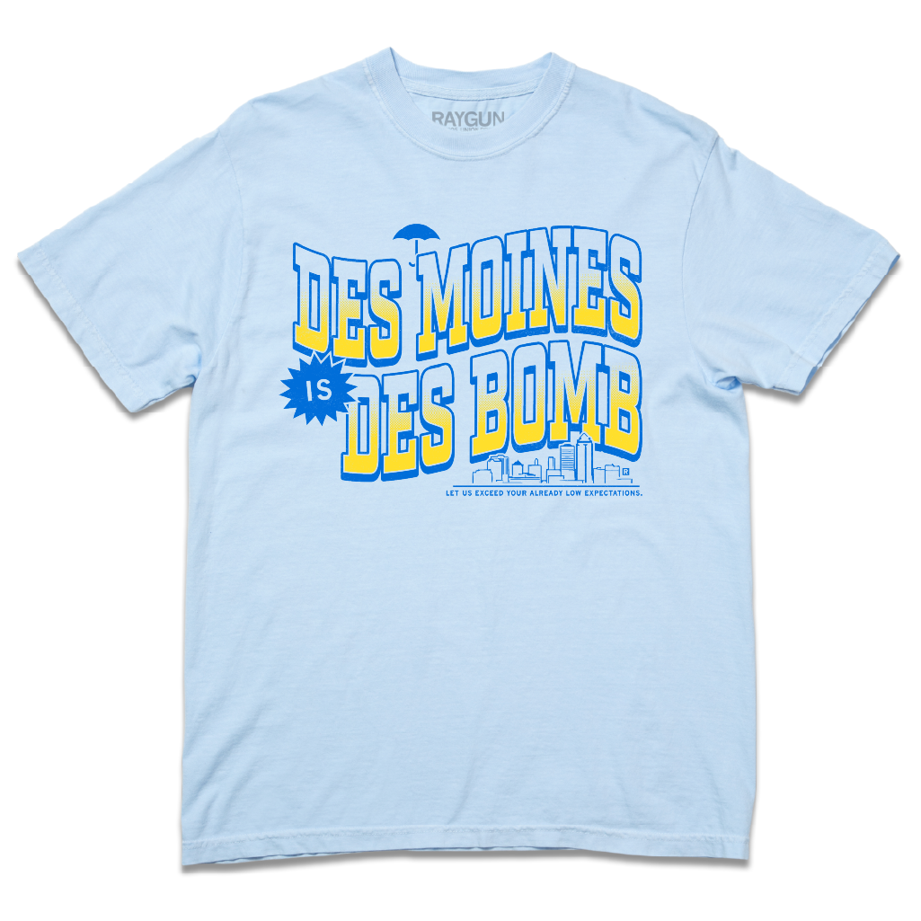 Comfort Colors heavyweight light blue t-shirt with bright gold and blue ink text that reads "Des Moines is Des Bomb. Let us exceed your already low expectations" with a small Des Moines skyline in the bottom right.
