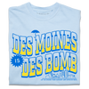 A folded heavyweight light blue t-shirt with bright gold and blue ink text that reads "Des Moines is Des Bomb. Let us exceed your already low expectations" with a small Des Moines skyline in the bottom right.