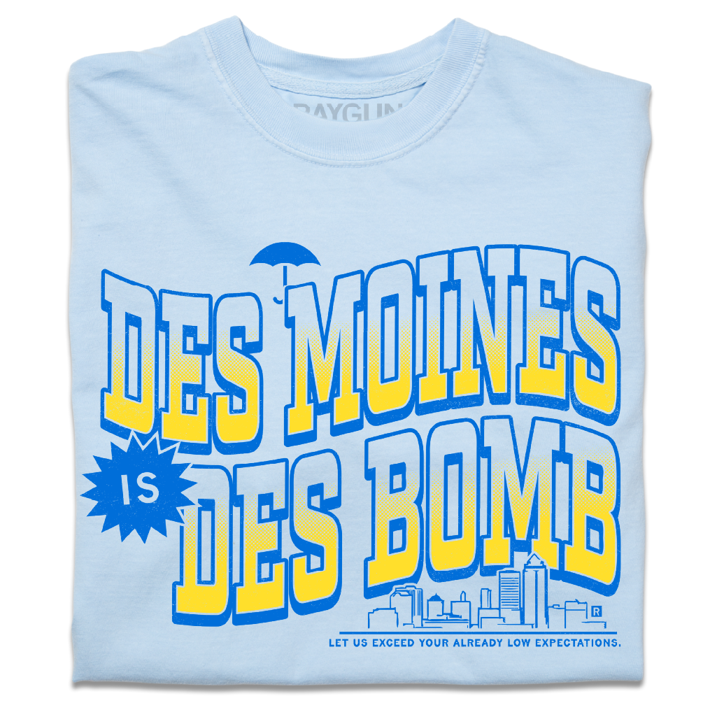 A folded heavyweight light blue t-shirt with bright gold and blue ink text that reads "Des Moines is Des Bomb. Let us exceed your already low expectations" with a small Des Moines skyline in the bottom right.