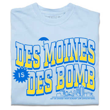 A folded heavyweight light blue t-shirt with bright gold and blue ink text that reads "Des Moines is Des Bomb. Let us exceed your already low expectations" with a small Des Moines skyline in the bottom right.
