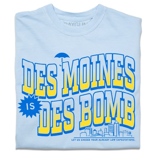 A folded heavyweight light blue t-shirt with bright gold and blue ink text that reads "Des Moines is Des Bomb. Let us exceed your already low expectations" with a small Des Moines skyline in the bottom right.