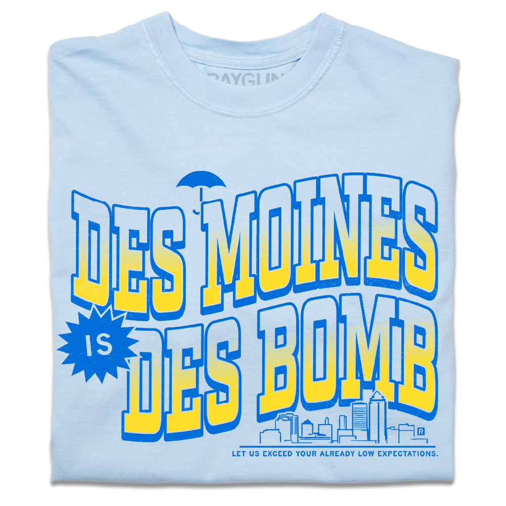 A folded heavyweight light blue t-shirt with bright gold and blue ink text that reads "Des Moines is Des Bomb. Let us exceed your already low expectations" with a small Des Moines skyline in the bottom right.