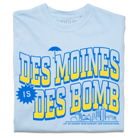 A folded heavyweight light blue t-shirt with bright gold and blue ink text that reads "Des Moines is Des Bomb. Let us exceed your already low expectations" with a small Des Moines skyline in the bottom right.