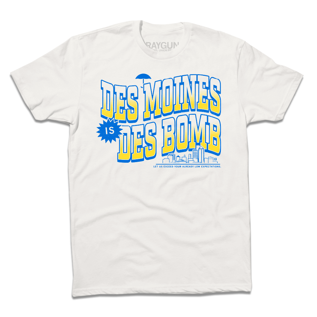 White t-shirt with bright gold and blue ink text that reads "Des Moines is Des Bomb. Let us exceed your already low expectations" with a small Des Moines skyline in the bottom right.
