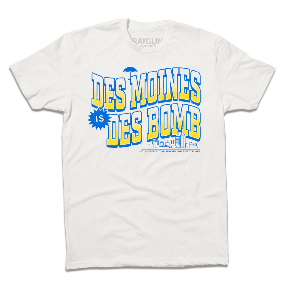 White t-shirt with bright gold and blue ink text that reads "Des Moines is Des Bomb. Let us exceed your already low expectations" with a small Des Moines skyline in the bottom right.