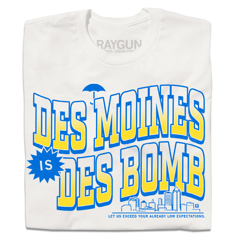 A folded off white t-shirt with bright gold and blue ink text that reads "Des Moines is Des Bomb. Let us exceed your already low expectations" with a small Des Moines skyline in the bottom right.