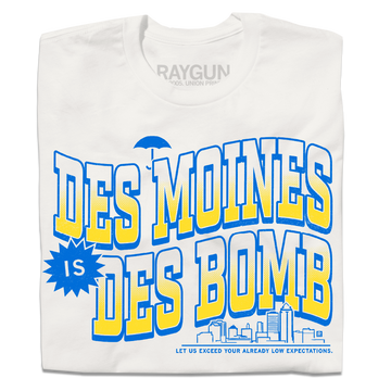 A folded off white t-shirt with bright gold and blue ink text that reads "Des Moines is Des Bomb. Let us exceed your already low expectations" with a small Des Moines skyline in the bottom right.