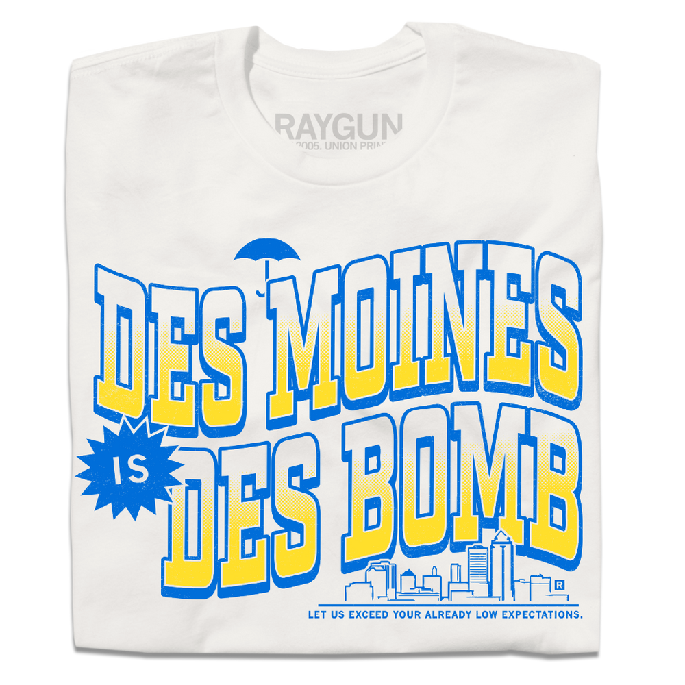 A folded off white t-shirt with bright gold and blue ink text that reads "Des Moines is Des Bomb. Let us exceed your already low expectations" with a small Des Moines skyline in the bottom right.