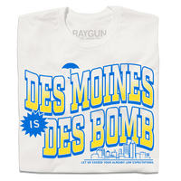 A folded off white t-shirt with bright gold and blue ink text that reads "Des Moines is Des Bomb. Let us exceed your already low expectations" with a small Des Moines skyline in the bottom right.