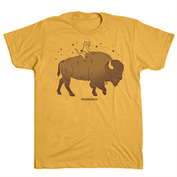 A dark gold t-shirt with a brown ink illustration of RAYGUN's cat mascot riding a bison over the text "Nebraska."
