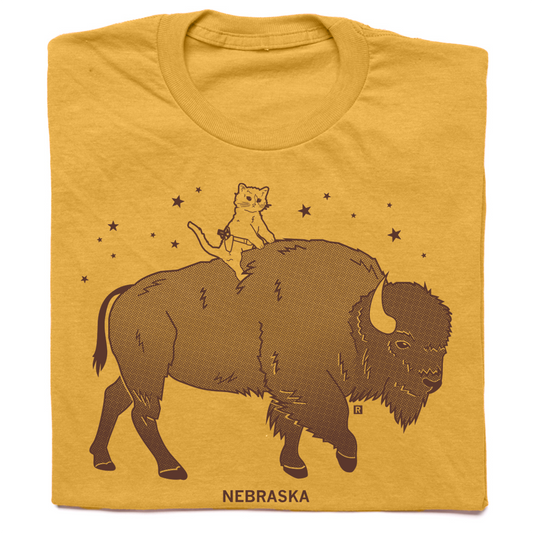 A dark yellow t-shirt with brown ink depicting RAYGUN's cat mascot riding a bison over the text "Nebraska."