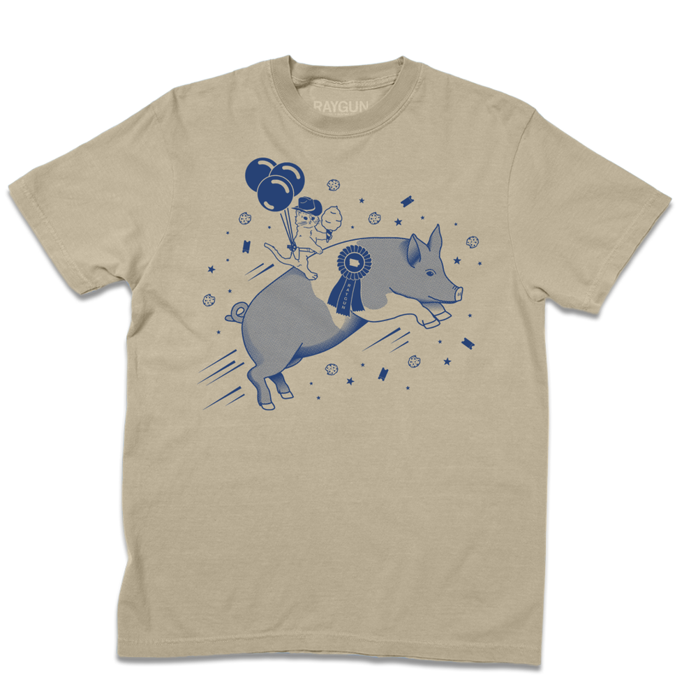 A folded khaki heavyweight t-shirt with royal blue print depicting a hat with a cowboy hat holding cotton candy while riding a giant pig. The cat is also wearing a holstered RAYGUN with three balloon attached to it.