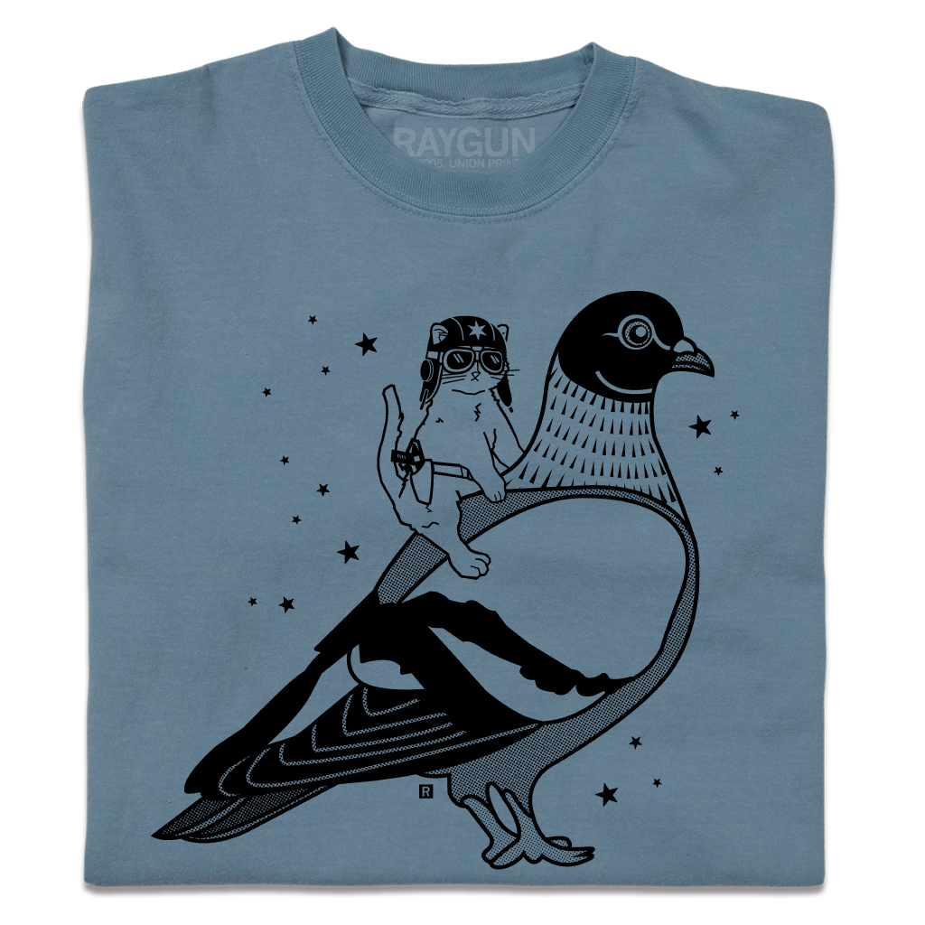 A folded denim blue heavyweight t-shirt with black ink print illustrating RAYGUN's cat mascot riding a giant pigeon.