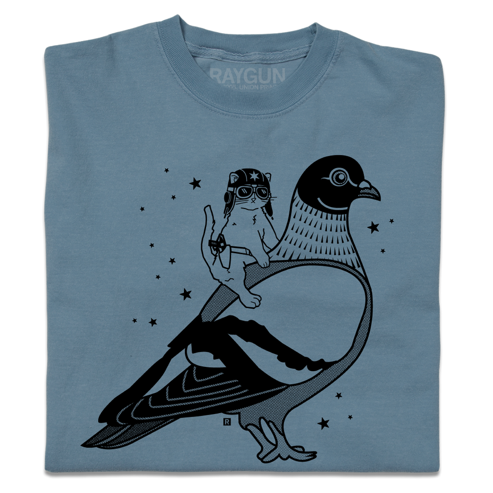 A folded denim blue heavyweight t-shirt with black ink print illustrating RAYGUN's cat mascot riding a giant pigeon.