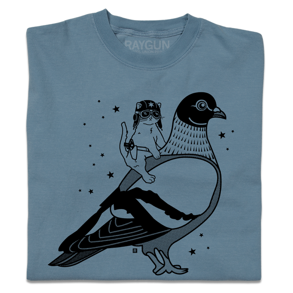 A folded denim blue heavyweight t-shirt with black ink print illustrating RAYGUN's cat mascot riding a giant pigeon.