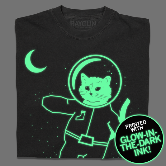 A folded dark grey shirt with glow in the dark ink depicting RAYGUN's cat mascot, Gary, as an astronaut with tiny stars and the moon behind him.