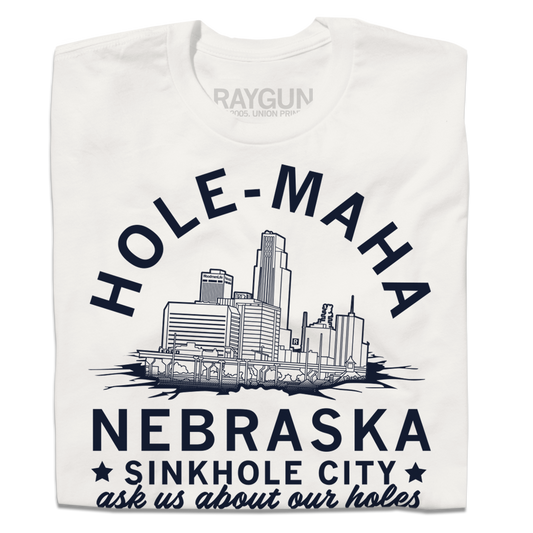 A folded off-white t-shirt with navy ink depicting downtown Omaha in a sinkhole with text that reads"Hole-maha, Nebraska. Sinkhole City, USA. As us about our holes."