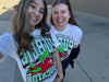 Two smiling women wearing white union labor t-shirts featuring a graphic of a green worm reading a book on a red apple with the word EDUCATOR in bold green letters.