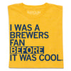 A folded heather gold t-shirt with left-aligned navy print that says, "I WAS A BREWERS FAN BEFORE IT WAS COOL."