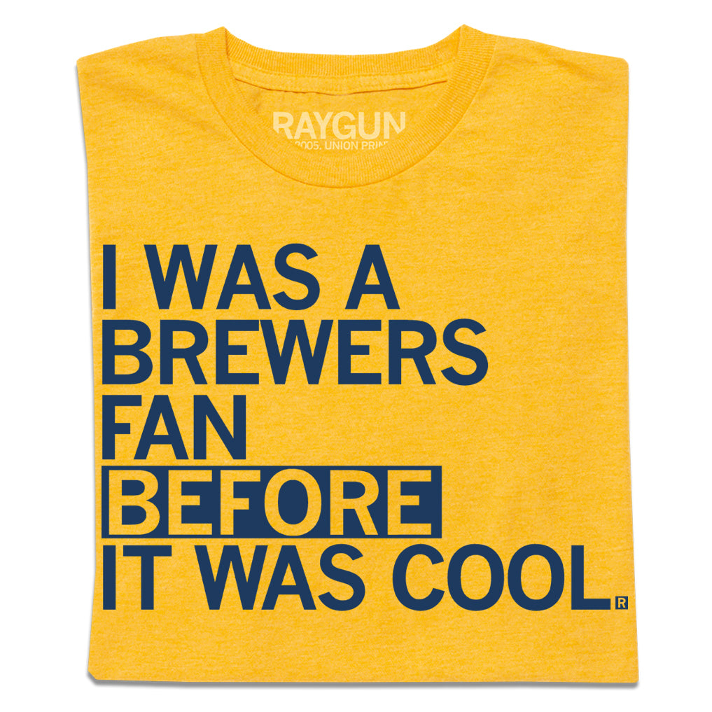 A folded heather gold t-shirt with left-aligned navy print that says, "I WAS A BREWERS FAN BEFORE IT WAS COOL."