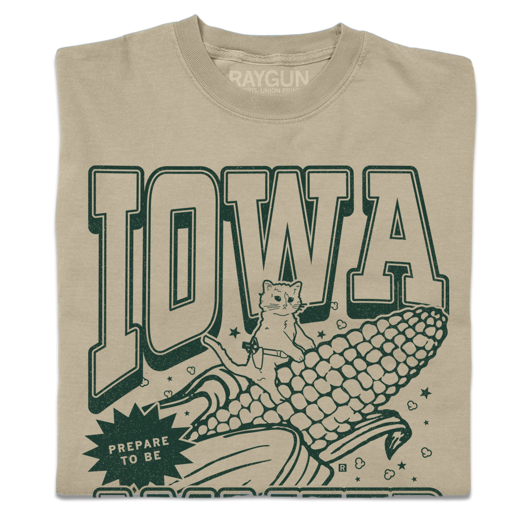 Tan garment-dyed t-shirt featuring a dark green graphic of a cat riding a giant ear of Iowa corn with the text IOWA and PREPARE TO BE.